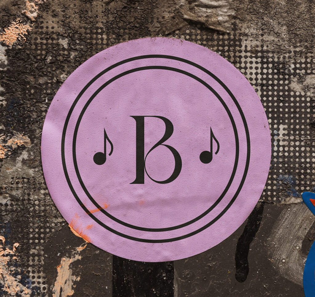 Ein blasslila runder Sticker ist auf einer urbanen Wand zu sehen. Darauf ist das "B" aus dem Logo von Beats and Bites und daneben jeweils eine kleine Note zu sehen. Das ganze wird umrahmt von zwei schwarzen Konturen eines Kreises.